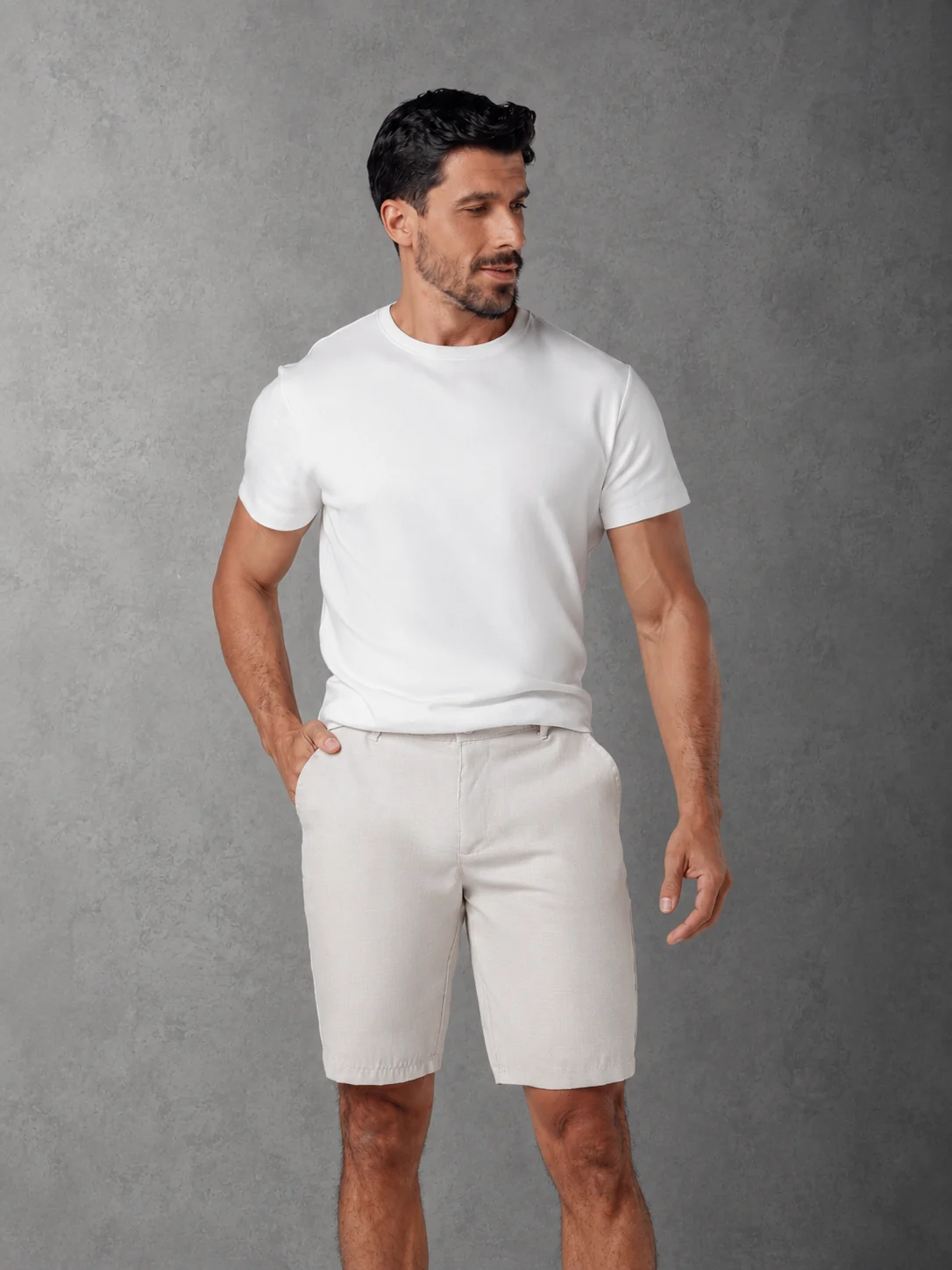 Modelo com bermuda clara e camiseta branca, diante de um fundo cinza