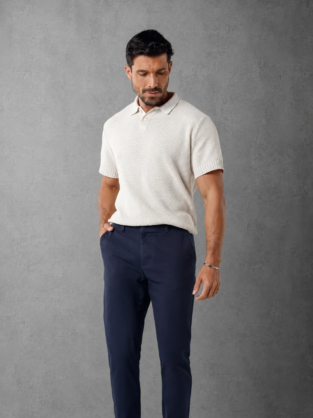 Modelo com calça casual azul-marinho e polo clara, diante de um fundo cinza